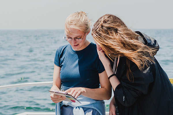 Young woman on a boat points at clipboard while another woman looks over her shoulder.