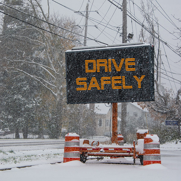 Electric road traffic mobile sign by the side of a snow covered road with snow falling that says, Drive Safely.